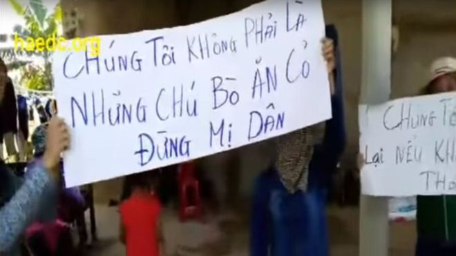 Người biểu tình giơ cao biểu ngữ trước cửa nhà trưởng thôn Cồn Sẻ - hình từ video clip đăng trên YouTube